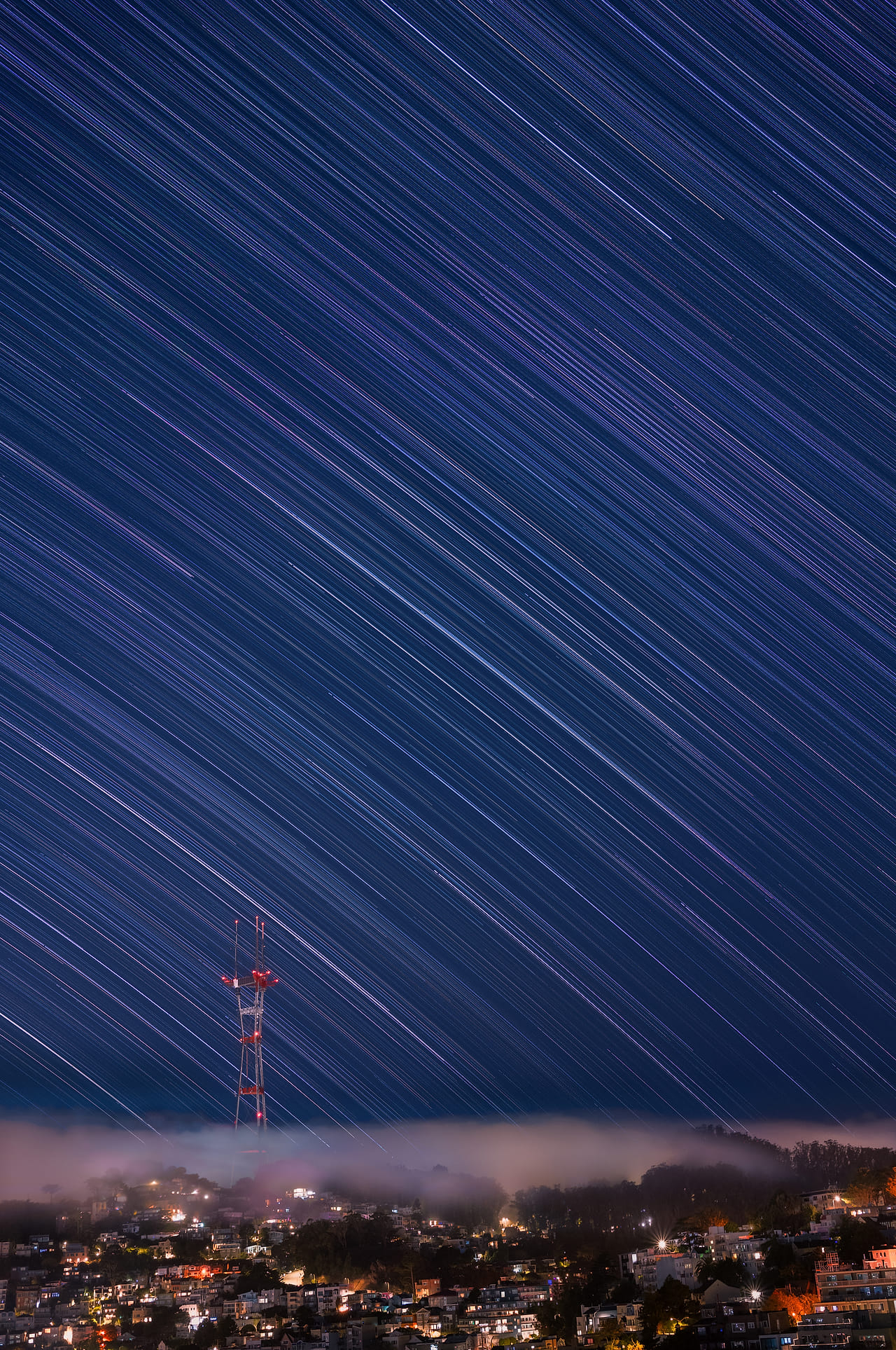 Long exposure of Sutro Tower at night, star trails streaking across the sky, with summer fog lying over the lit-up houses below Corona Heights.