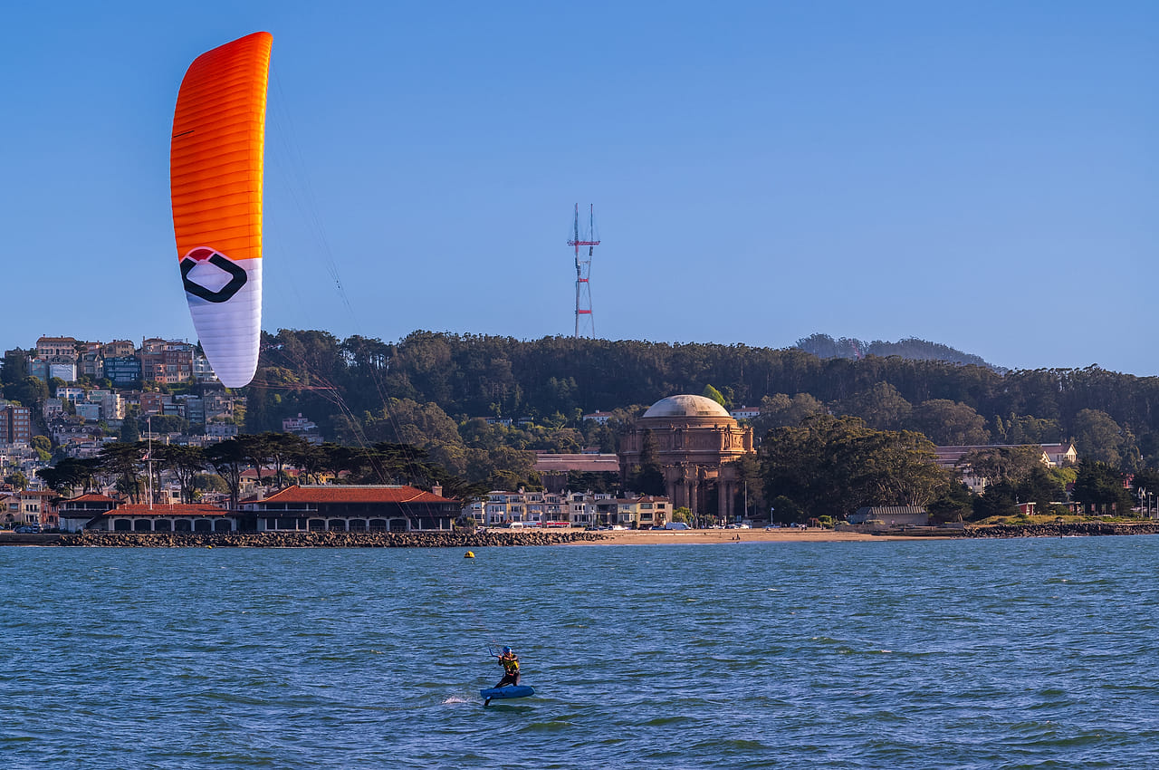 A kitesurfer under an orange kite on the bay off the Marina, the Palace of Fine Arts on the shore and Sutro Tower on the hill behind.