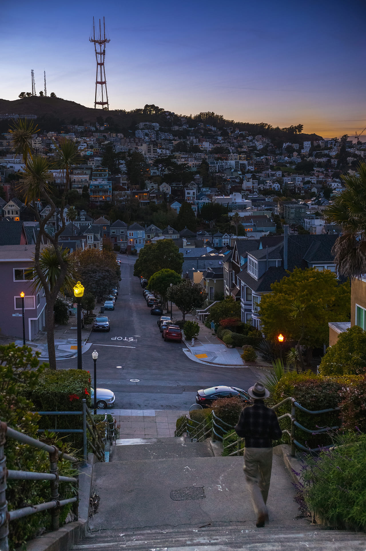 A man in a hat walking down the Liberty Street steps at dusk, the street dropping away below him and Sutro Tower on the ridge against an orange sky.